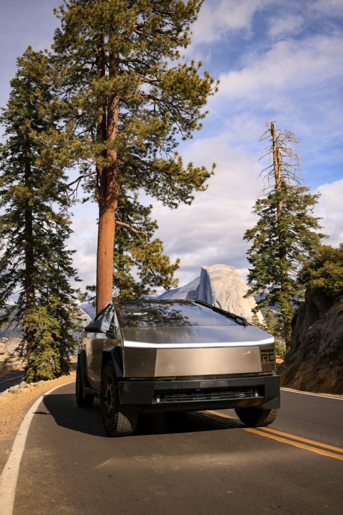 A Tesla Cybertruck drives along a scenic mountain road with towering pine trees and clear skies.